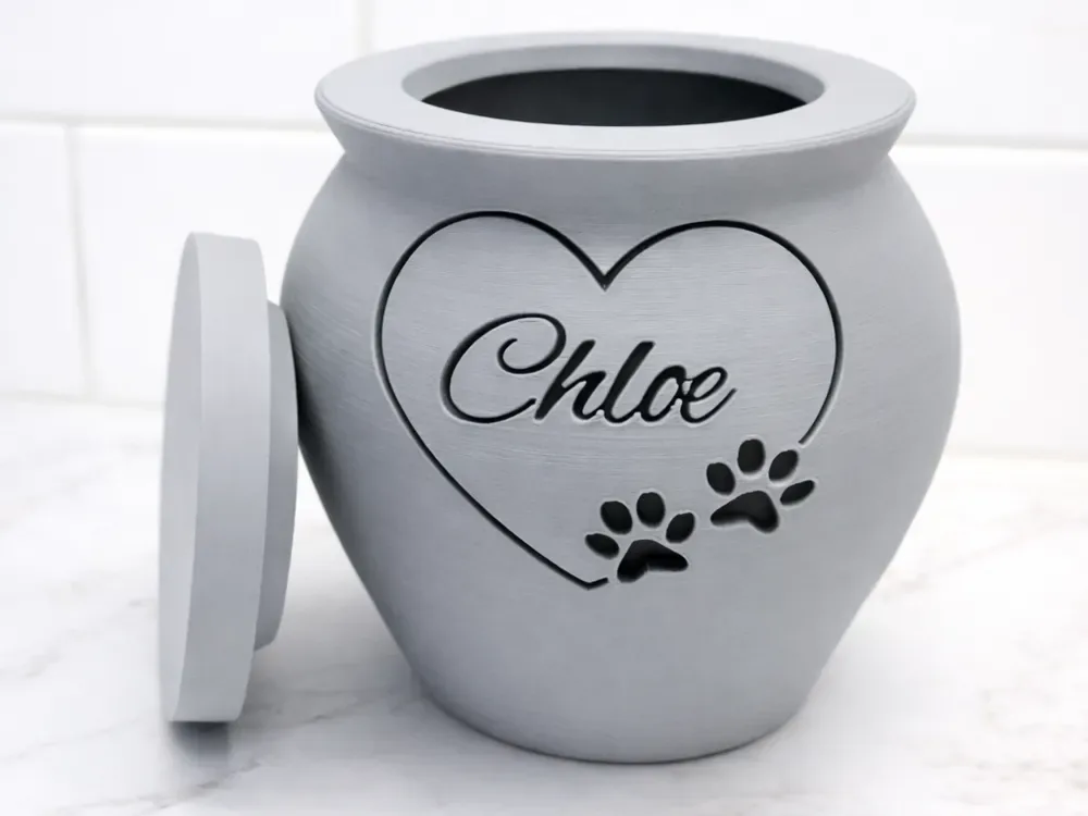 Urna pet cinza em formato de vaso, com o nome do pet gravado dentro de um coração e duas marcas de pata, tampa ao lado